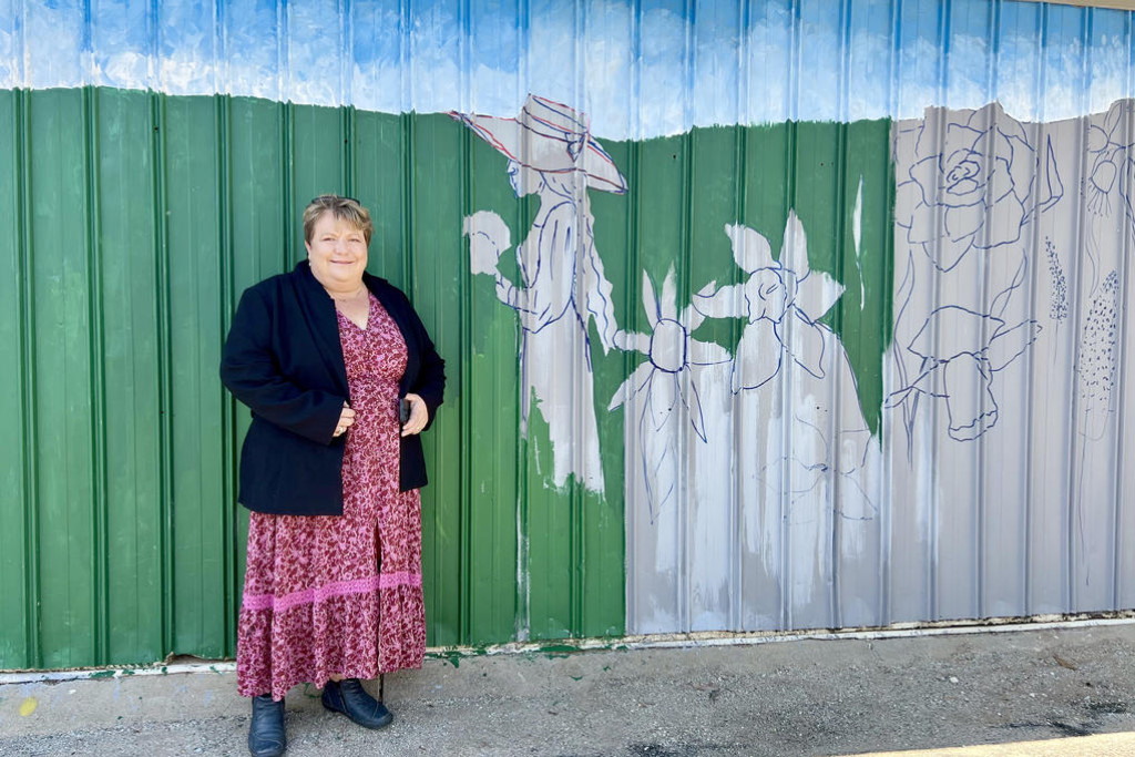 Belinda Eckermann stands in front of the mural-in-progress at the Old Shelter Shed, inviting the community to help bring the vibrant garden scene to life.